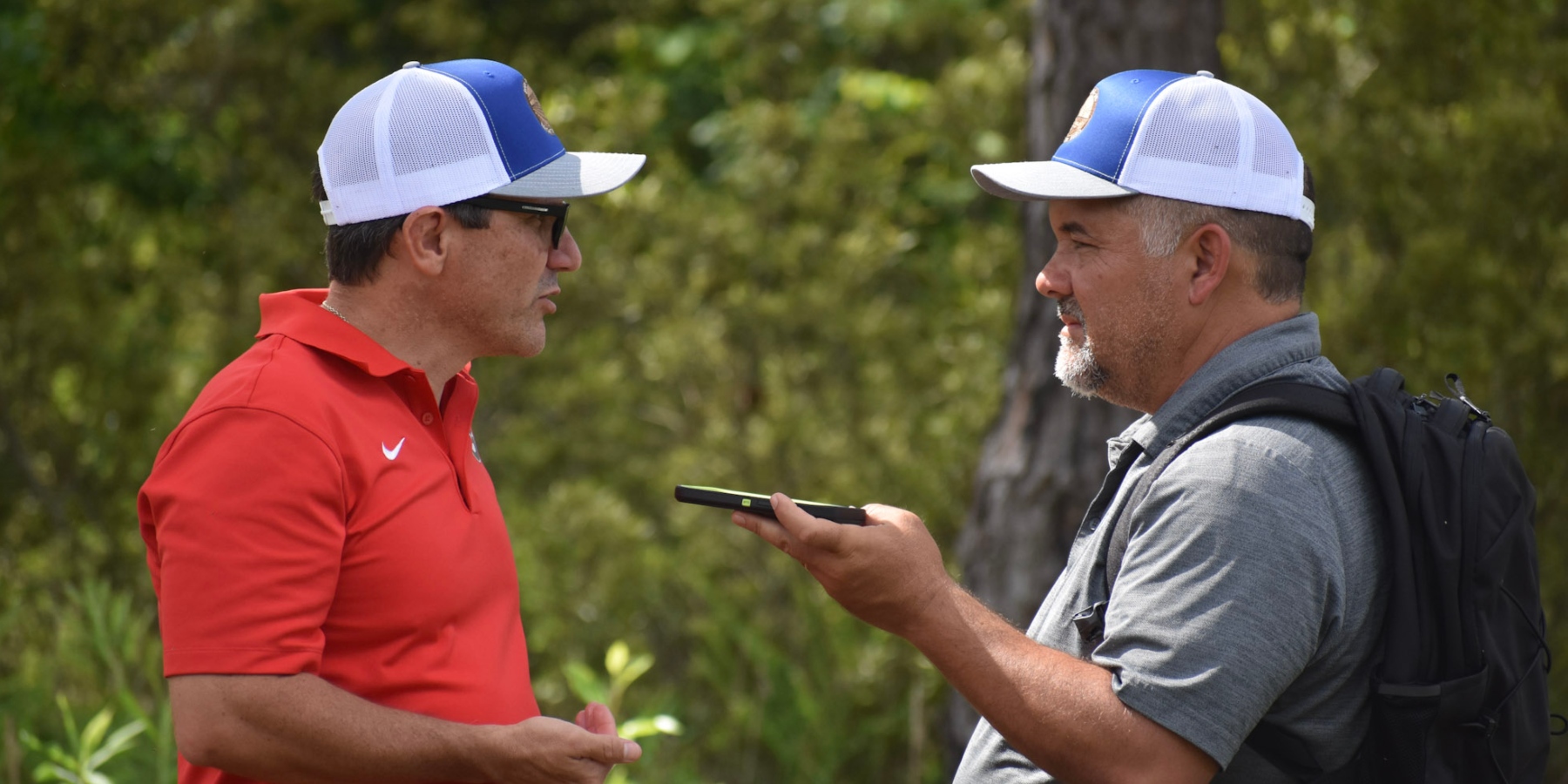 Two people (Dr. Ted McAvoy, left and Clint Thompson, right) stand outdoors in a wooded setting conducting an interview.